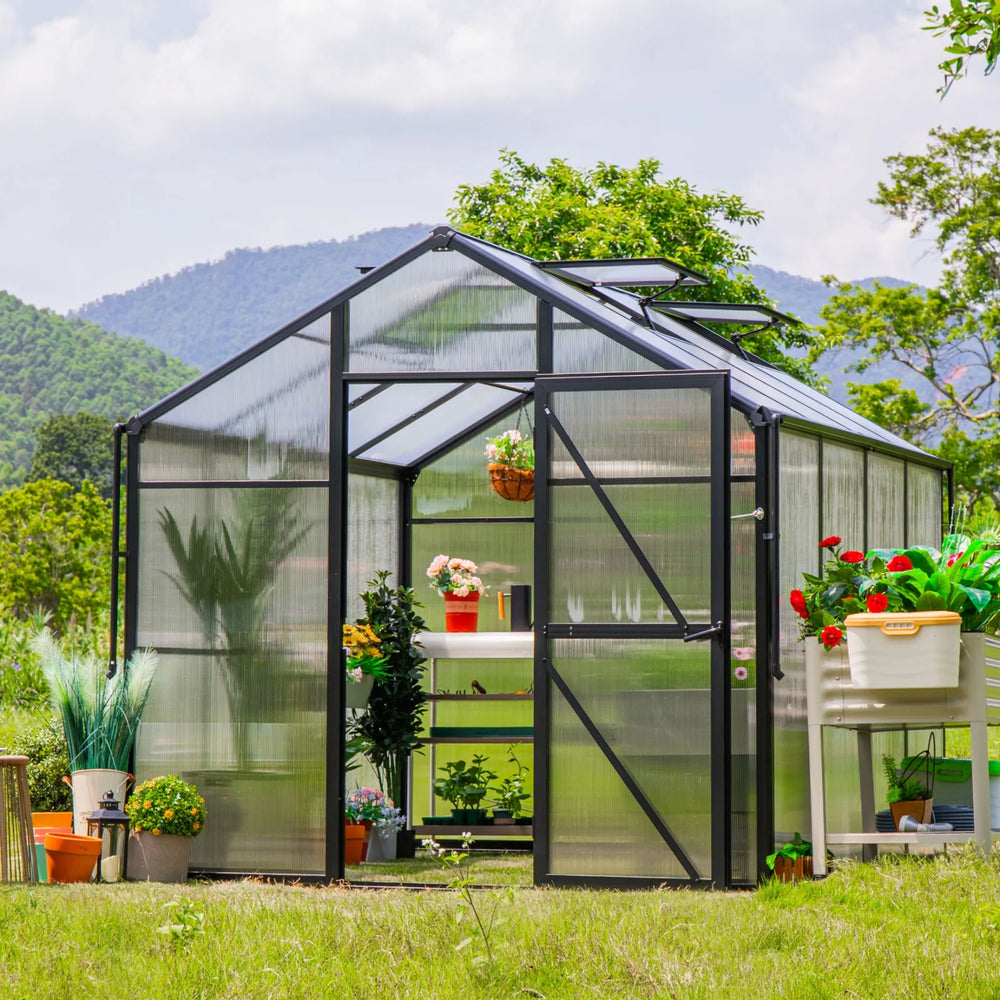 Vego Junior Victorian Greenhouse 8ft wide in a landscaped backyard with open hinged door, roof vents, potted plants, and gardening accessories